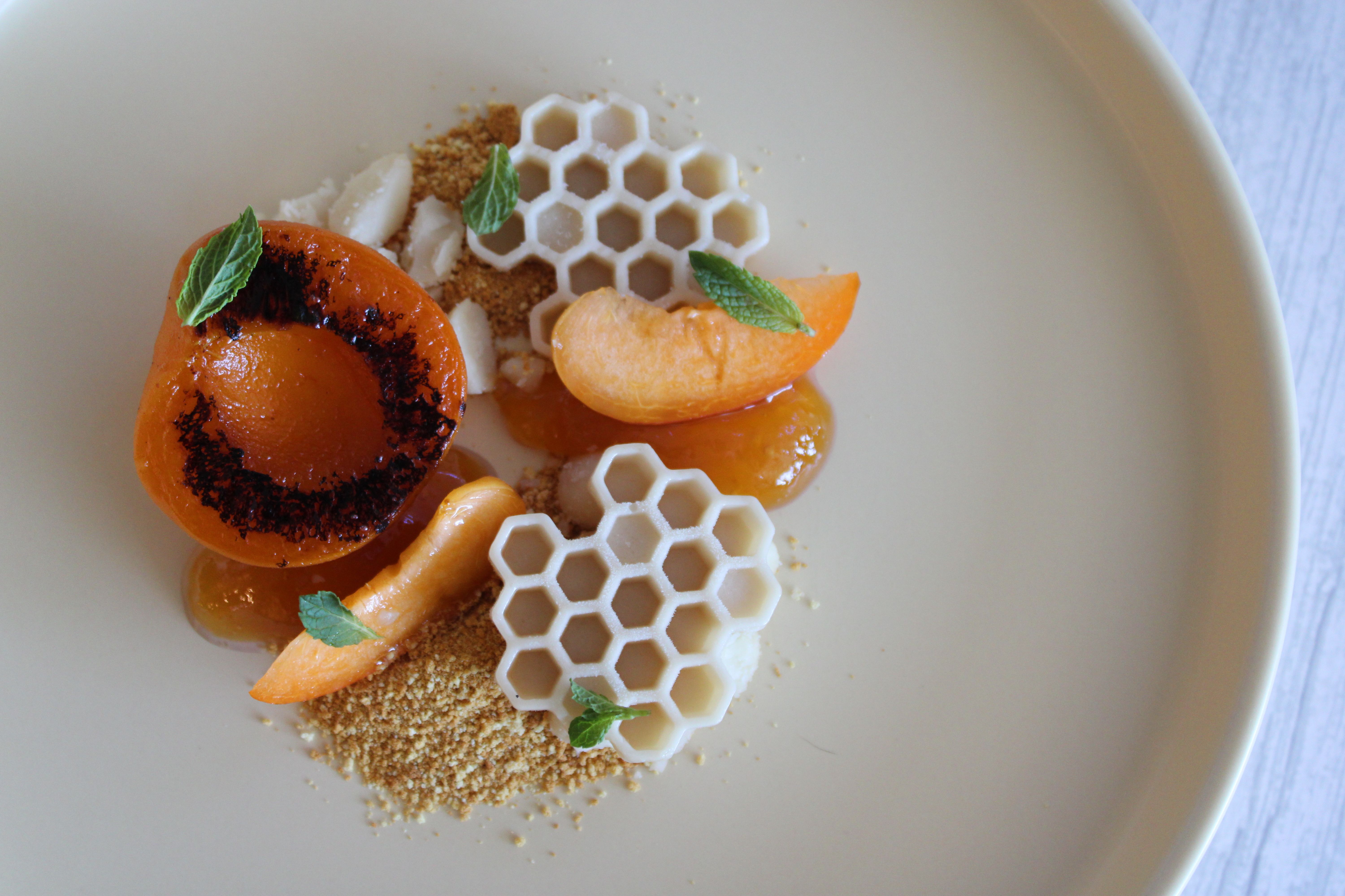 Gourmet apricot dessert with a grilled apricot half and slices, white honeycomb-shaped chocolate, golden biscuit crumble, apricot gel and fresh mint leaves on a beige plate.