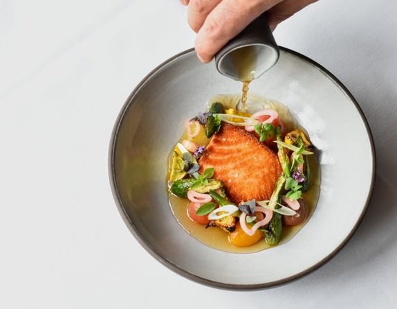 Top-down view of a seared salmon fillet in a modern ceramic bowl surrounded by asparagus, blistered tomatoes, pickled onions and microgreens as a hand pours a light broth — fine-dining seafood plate.