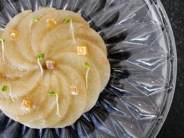 Top-down close-up of a pale swirled molded dessert on a faceted glass plate, garnished with tiny green microgreens and small candied citrus cubes