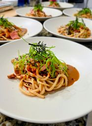 Udon-style noodles in savory sauce topped with shredded nori, scallion curls, microgreens and sesame seeds, plated in white bowls on a restaurant prep counter.