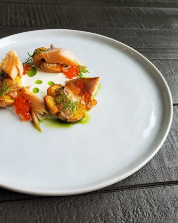 Gourmet plated appetizer on a large white plate: smoked salmon and seared baby potatoes topped with orange salmon roe, dill sprigs and drops of green herb oil on a dark wooden table.