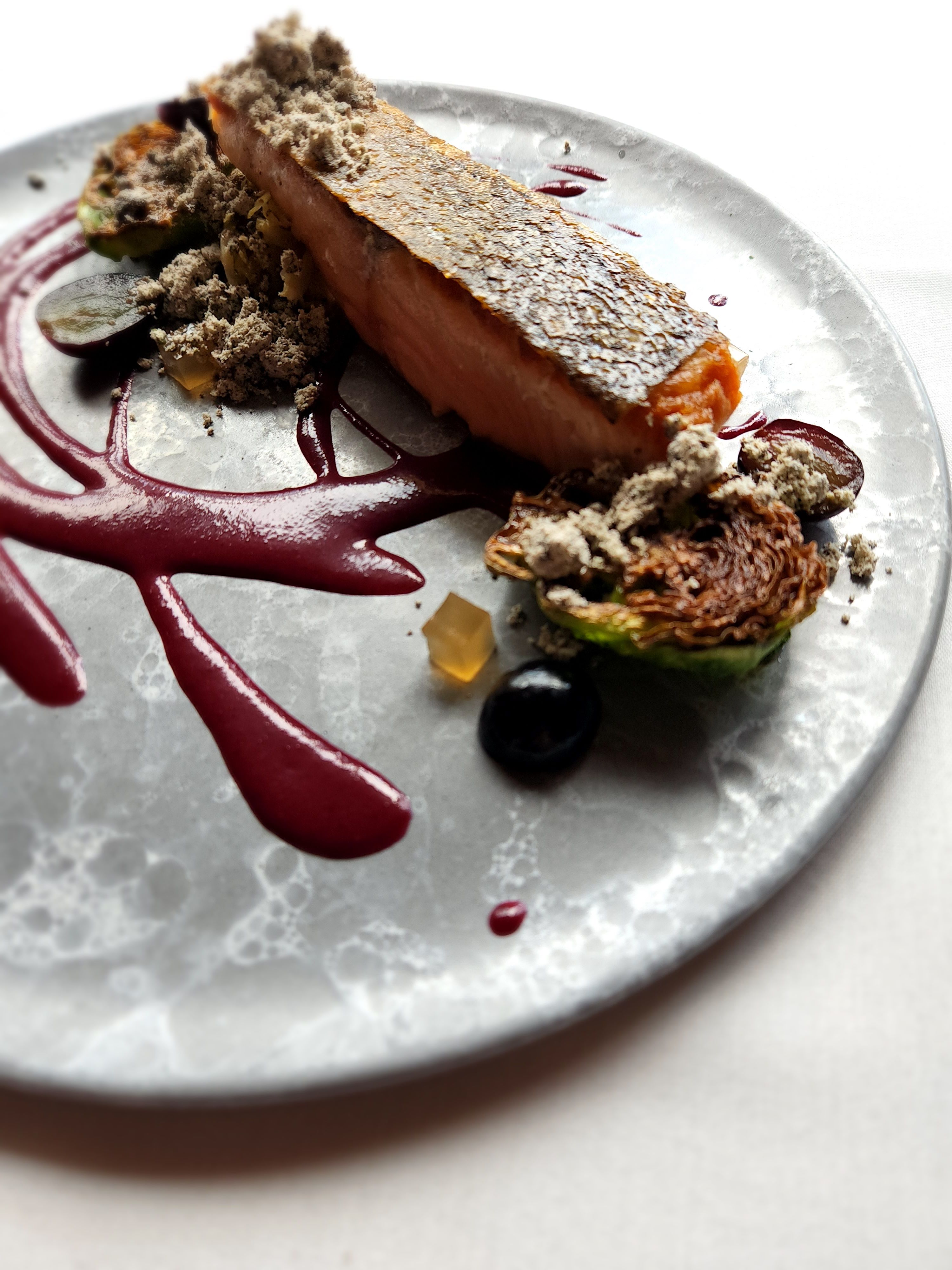 Seared salmon fillet with crispy skin on a gray plate, red berry purée swirls, roasted Brussels sprout, grapes and crumb garnish — modern fine-dining plating.