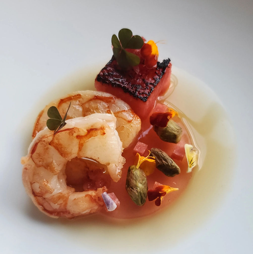 Seared shrimp and charred watermelon cube garnished with pistachios, microgreens and edible flowers in a light citrus broth on a white plate — elegant seafood appetizer.