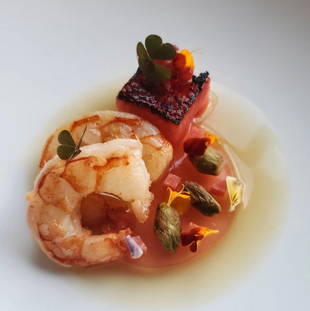 Seared shrimp and charred watermelon cube garnished with pistachios, microgreens and edible flowers in a light citrus broth on a white plate — elegant seafood appetizer.