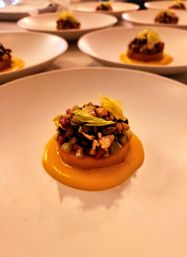Fine-dining appetizer: a mini savory tartlet on a bright orange squash purée, topped with diced vegetables, seeds and nuts and a celery leaf, served on a white ceramic plate with more plated servings blurred in the background.