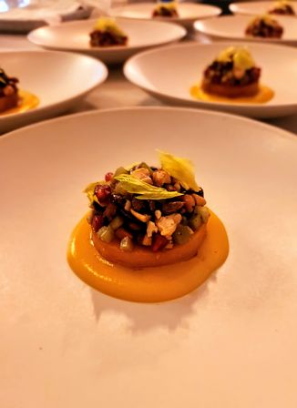 Fine-dining appetizer: a mini savory tartlet on a bright orange squash purée, topped with diced vegetables, seeds and nuts and a celery leaf, served on a white ceramic plate with more plated servings blurred in the background.