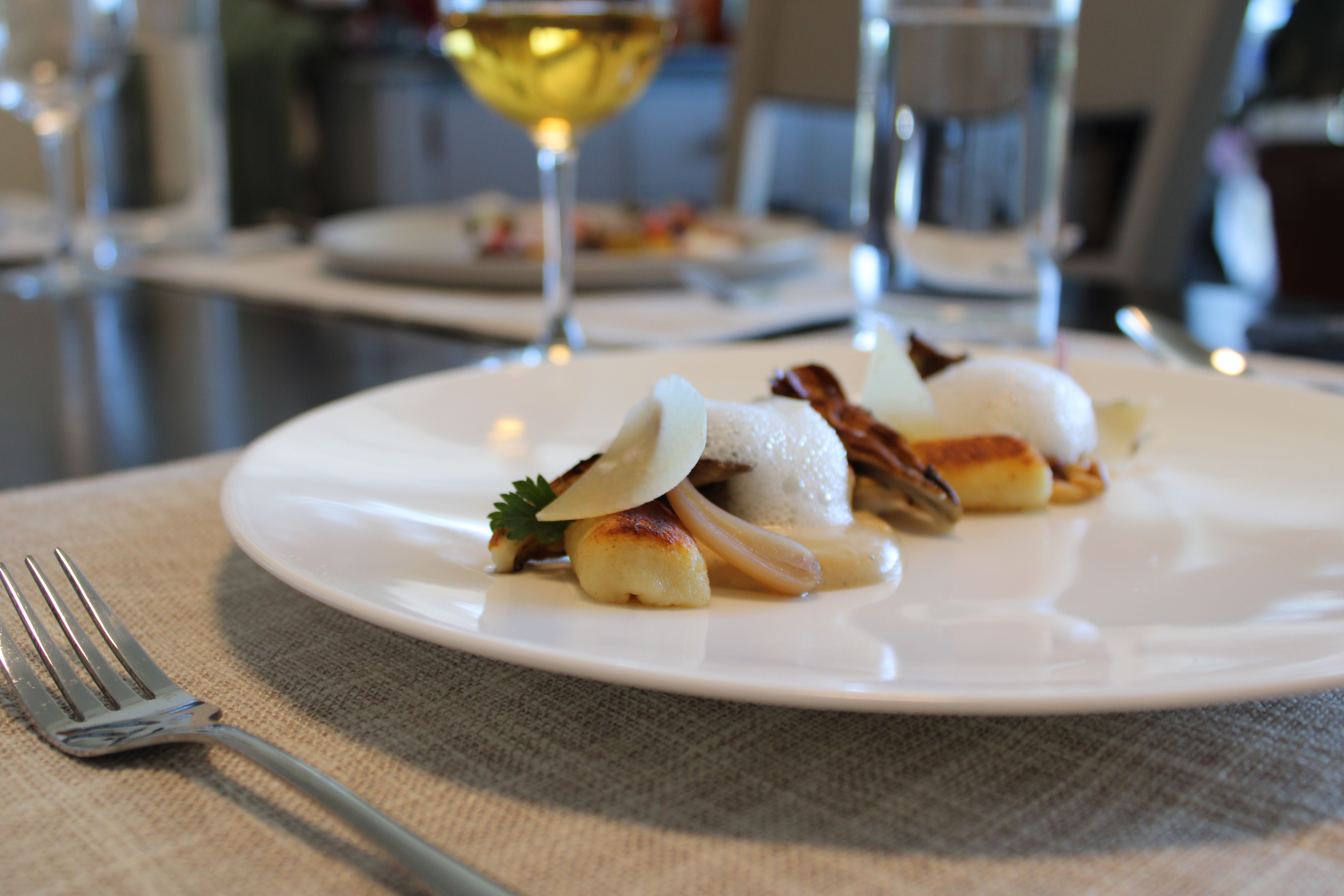 Pan-seared gnocchi with mushrooms, foam and shaved cheese elegantly plated on a white dish in a fine-dining restaurant, fork on a linen placemat and a glass of white wine blurred in the background.