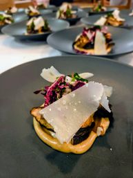 Close-up of a gourmet appetizer on a matte black plate — roasted mushroom on a creamy purée topped with shaved hard cheese and vibrant purple cabbage slaw, multiple plated servings blurred in the background.