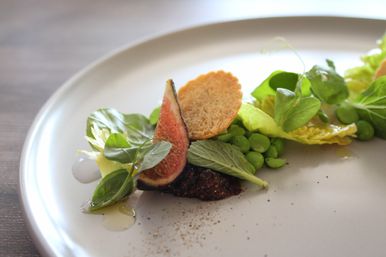Minimalist fine-dining salad on a white plate: halved fresh fig, bright green peas and pea shoots, baby romaine leaves, a round crisp crostini, dollop of fig jam and olive oil drizzle.