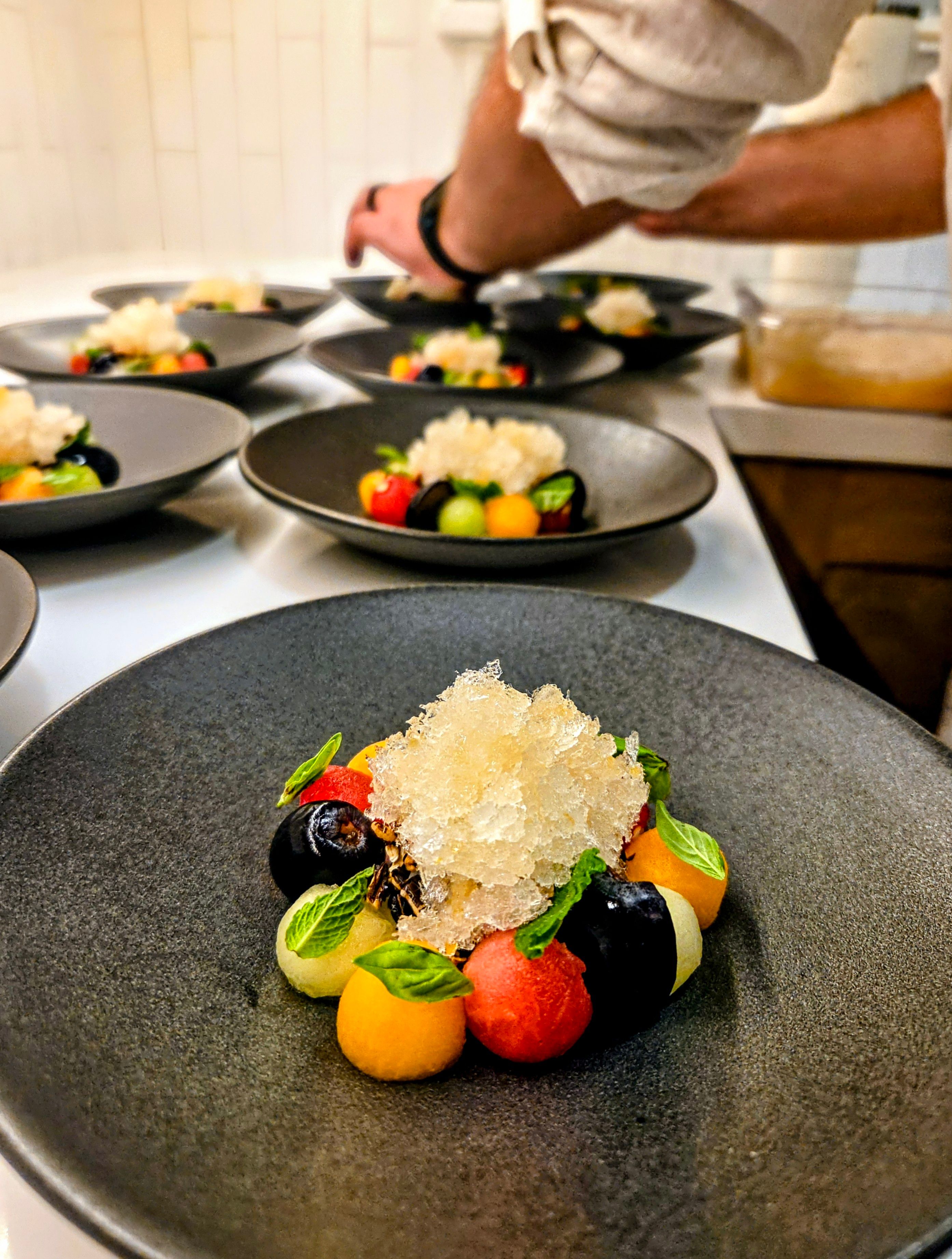 Close-up of a gourmet plated fruit dessert on a black ceramic plate — colorful melon and grape spheres with fresh mint leaves topped with sparkling granita, chef’s hands arranging multiple dishes in a restaurant kitchen.