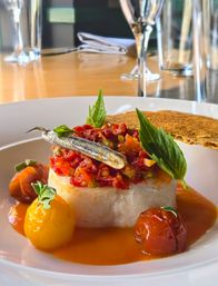 Gourmet Mediterranean-style seafood appetizer — creamy white fish cake topped with chopped tomato ragout and a whole sardine, fresh basil leaves, blistered cherry tomatoes, orange tomato sauce and a crisp cracker on a white plate in a fine-dining setting.
