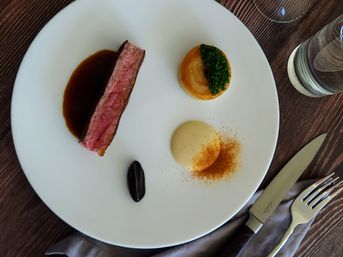 Top-down view of a gourmet plated steak course: medium-rare strip with demi-glace, herb-topped potato stack, creamy purée dusted with spice and a glossy sauce drop on a white plate, with knife, fork and water glass on a wood table — fine dining presentation.