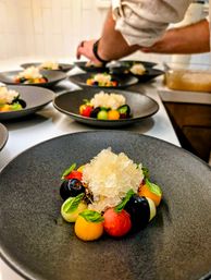 Fine-dining dessert on a matte black plate — colorful melon and fruit spheres with fresh herb leaves arranged around a central mound of sparkling granita, chef's hands plating multiple dishes at a busy kitchen station.