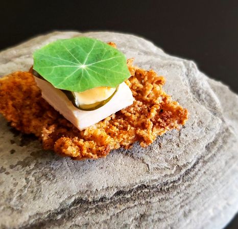 Modern gourmet appetizer on a textured stone slab — crunchy fried crisp topped with a square of soft cheese or tofu, cucumber slice, creamy dollop and bright nasturtium leaf.