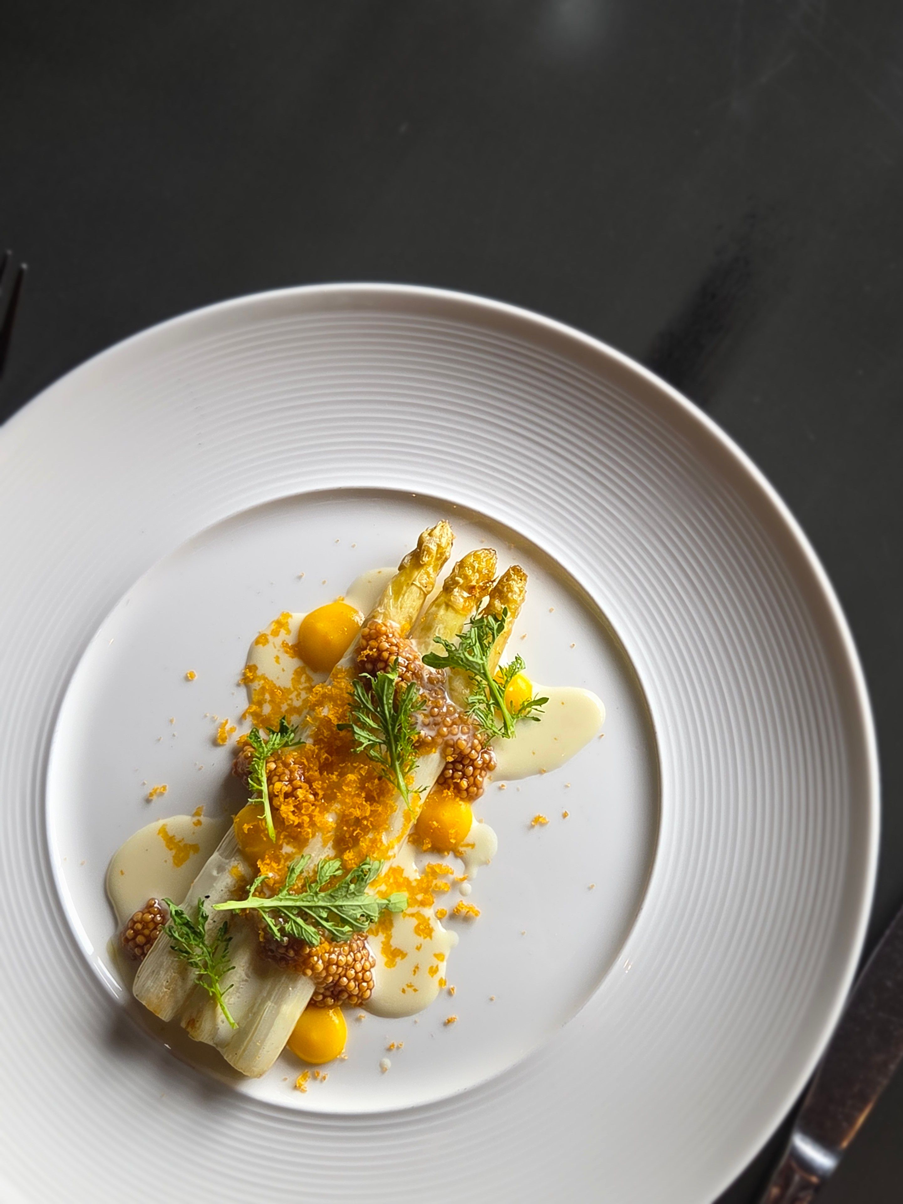 Gourmet white asparagus spears with whole-grain mustard pearls, bright yellow sauce dots, creamy emulsion, crunchy crumbs and microgreens artfully plated on a white rimmed dish — fine-dining seasonal spring plate on a dark table.