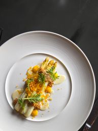 Gourmet white asparagus spears with whole-grain mustard pearls, bright yellow sauce dots, creamy emulsion, crunchy crumbs and microgreens artfully plated on a white rimmed dish — fine-dining seasonal spring plate on a dark table.