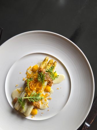 Gourmet white asparagus spears with whole-grain mustard pearls, bright yellow sauce dots, creamy emulsion, crunchy crumbs and microgreens artfully plated on a white rimmed dish — fine-dining seasonal spring plate on a dark table.