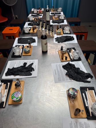 Long stainless-steel table set for a DIY fragrance‑blending workshop: multiple place settings with small glass vials, spray bottles, colorful sample tins, black nitrile gloves, wooden trays and instruction sheets, colorful stools and teal curtain in the background.
