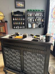 Black wooden counter with metal stamping tools on top, wall shelves of vintage apothecary bottles and green glassware, decorative skull and stained-glass window in a boutique interior.