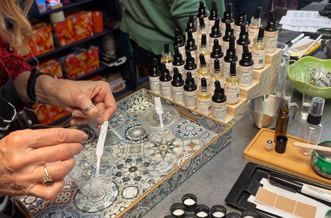 Hands testing fragrance strips at an artisan perfume-making station with rows of dropper bottles, sample cups, and measuring tools on a decorative tray
