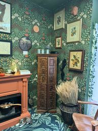 Cozy vintage-style corner with green botanical wallpaper, terracotta mantel, ornate carved wooden cabinet, framed fruit prints, wicker basket of dried grasses and a tabby cat curled by the hearth.