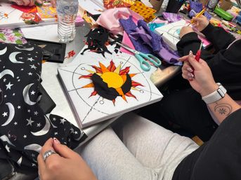 Close-up of hands creating a sun-and-moon fabric collage on a craft table, with scissors, colorful fabric scraps, star-patterned cloth, and a water bottle nearby