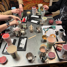 Overhead shot of a busy DIY candle-making workshop table with hands pouring colorful wax into molds, wooden wicks, small metal pouring cups, scissors and craft tools.