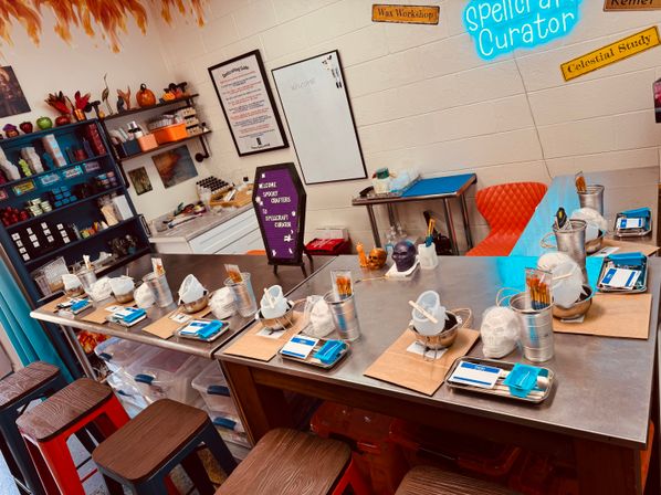 Spooky-themed craft studio with metal worktables set for a group mask-painting workshop — individual stations with plaster skull masks, paintbrushes, metal buckets, trays and name tags, shelves of supplies and Halloween decor on the walls.