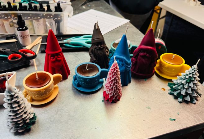 Colorful holiday novelty candles—gnome-shaped figures, teacup candles, and layered Christmas tree candles—arranged on a metal craft table with scissors, dye bottles, and candle-making supplies in the background.