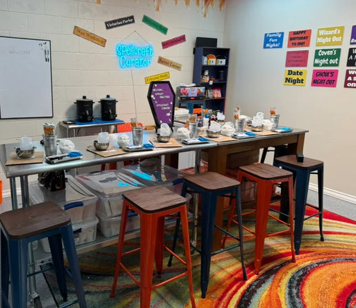 Colorful craft studio set for a spell-themed workshop: long tables with stools, mixing bowls, jars, tools and placemats, neon wall accent and themed signs, over a vivid circular rug.