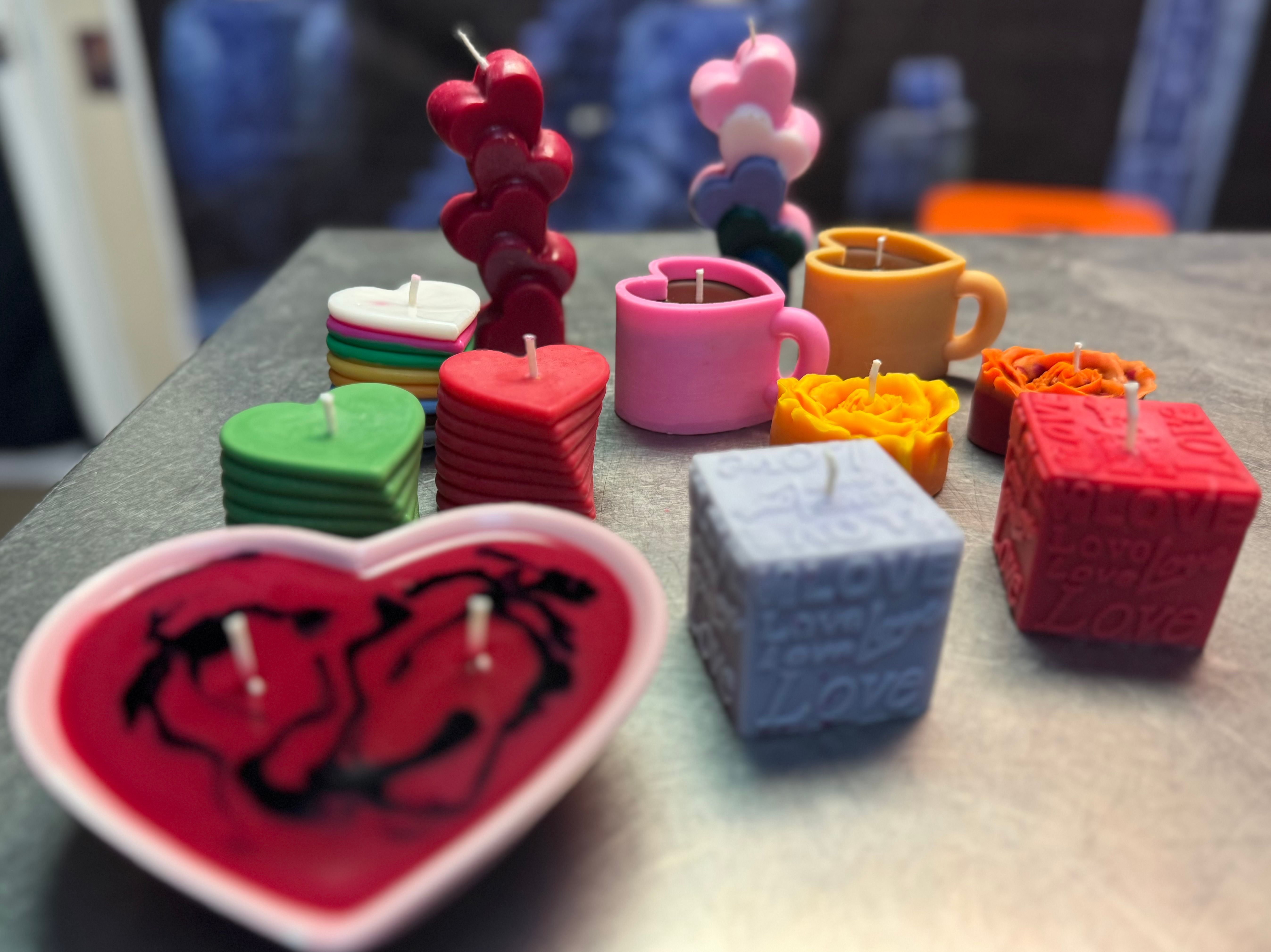 Bright, playful assortment of handmade candles on a tabletop: heart-shaped bowls and stacked heart disks, heart-on-a-stick stacks, mug-shaped candles, rose blooms, and embossed 'LOVE' cubes.