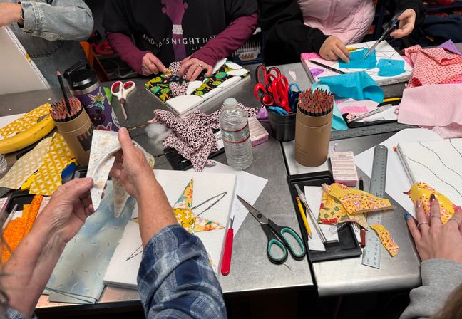 Busy craft workshop table with several hands cutting and arranging colorful patterned fabric pieces into collage shapes on small canvases, surrounded by scissors, colored pencils, rulers and a water bottle.