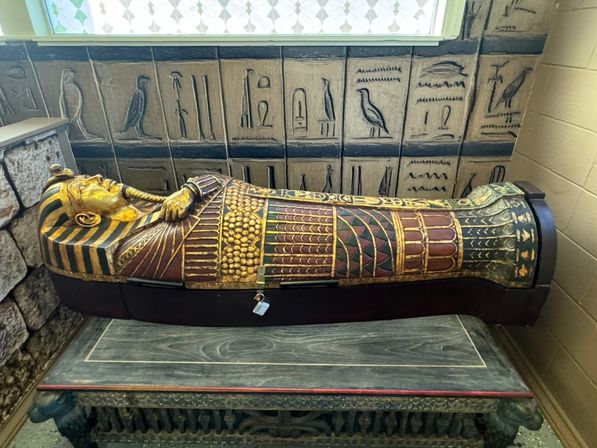 Ornate Egyptian-style sarcophagus replica with gold pharaoh mask and red-green patterned body resting on a carved wooden table against a tiled hieroglyphic wall in an indoor display.