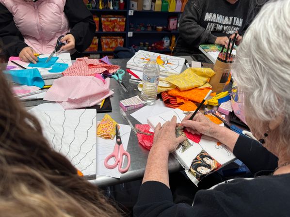 Busy community craft table with people cutting and gluing colorful fabric scraps onto small canvases, surrounded by scissors, colored pencils, patterned textiles and a water bottle.
