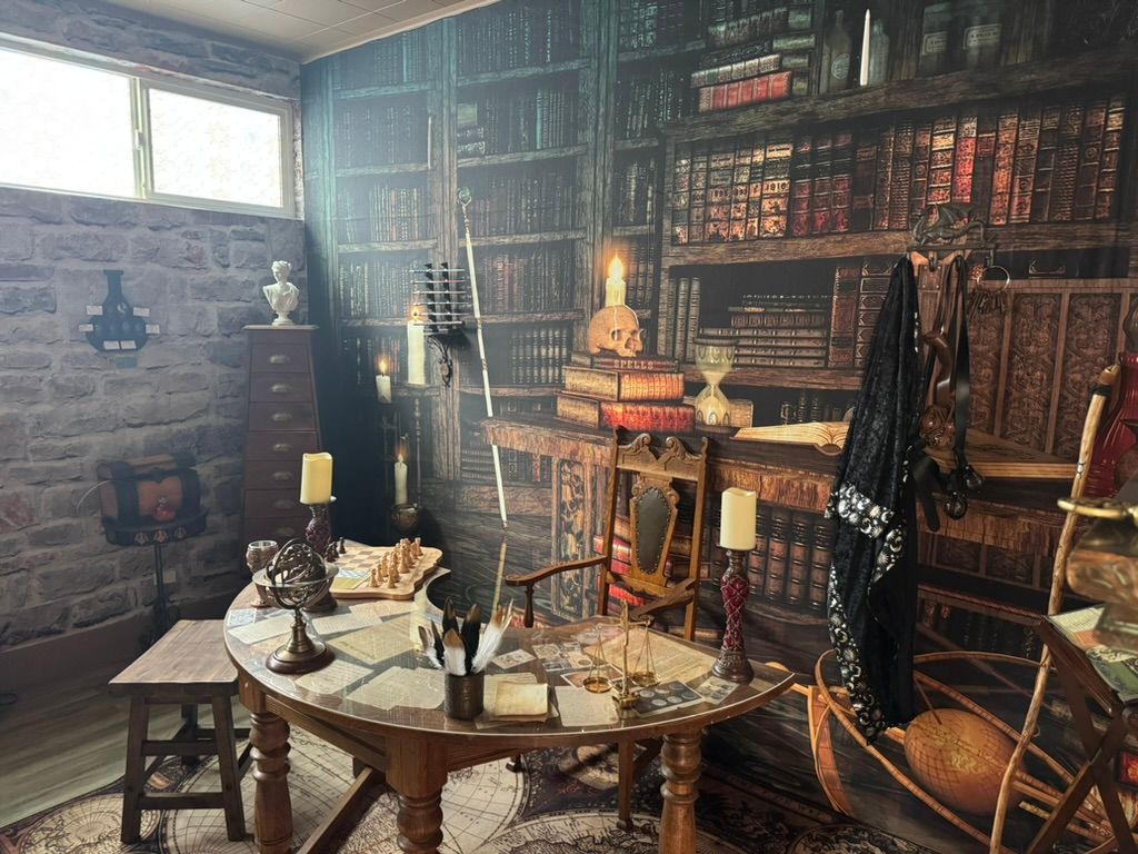 Cozy wizard study–themed basement room with stone walls and high window, faux floor-to-ceiling bookshelves, ornate wooden table laid with maps, quill, globe, hourglass, brass scales, candles and a chessboard, carved chair with skull candle and a black cloak draped nearby.