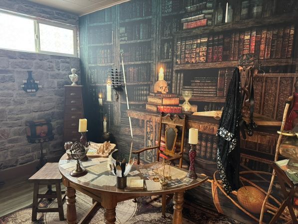 Cozy wizard study–themed basement room with stone walls and high window, faux floor-to-ceiling bookshelves, ornate wooden table laid with maps, quill, globe, hourglass, brass scales, candles and a chessboard, carved chair with skull candle and a black cloak draped nearby.