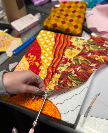 Close-up of a hand cutting bright red, yellow and orange patterned fabrics for a quilt on a craft table, with scissors, a foam template and sewing tools nearby.