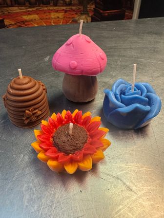 Colorful handmade decorative candles on a metal countertop: pink mushroom candle, blue rose candle, red-and-yellow sunflower candle, and a brown beehive-style candle.