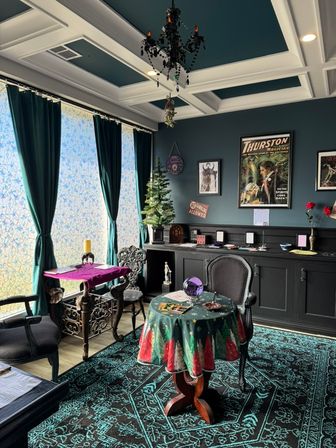 Cozy Victorian-style fortune-telling parlor with teal coffered ceiling and chandelier, velvet curtains, vintage magician posters on the wall, a small decorated tree, round table draped in a festive cloth holding a purple crystal ball and tarot cards, ornate chairs and a patterned rug.