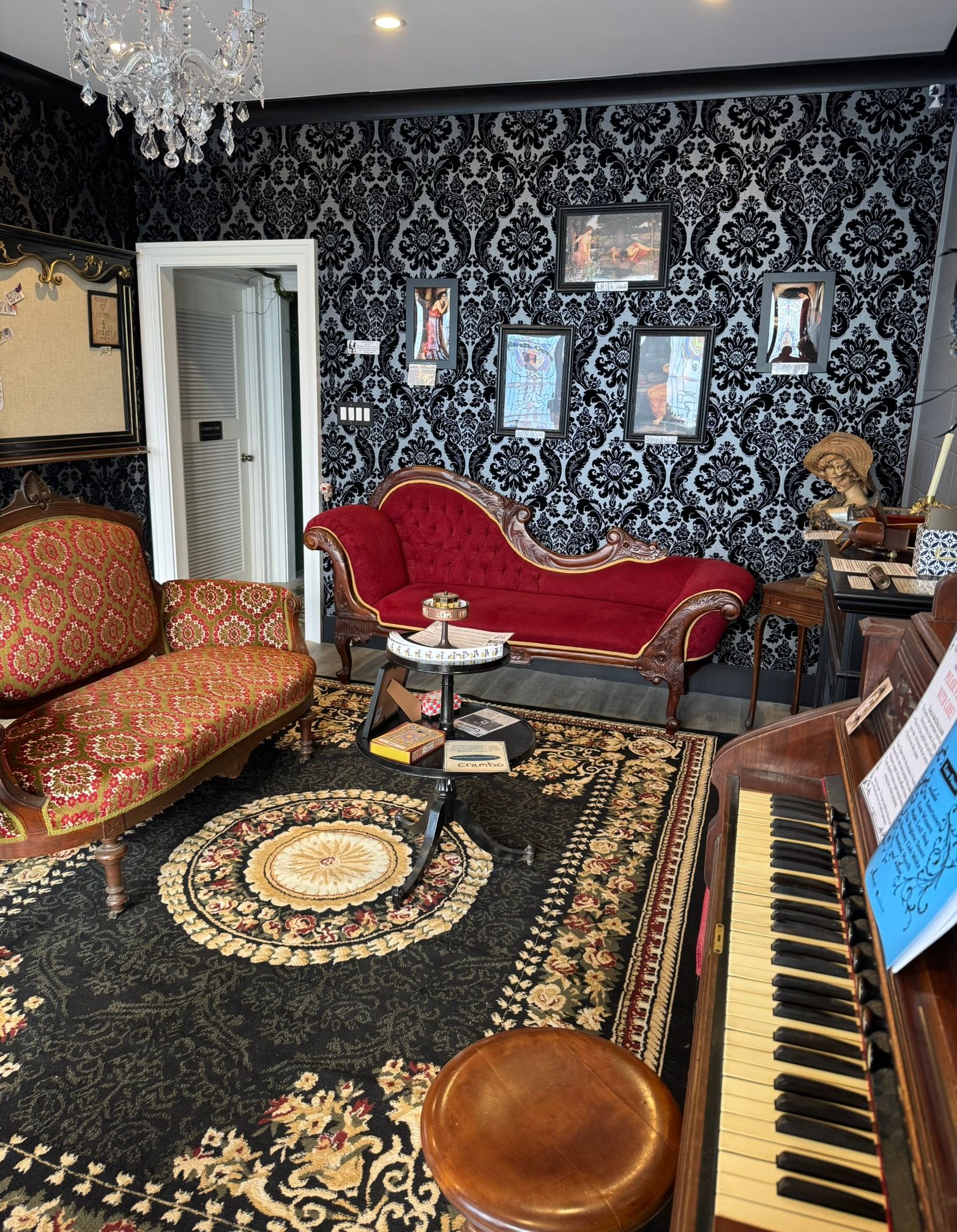 Victorian-style parlor interior with black damask wallpaper, red velvet chaise lounge and patterned settee, upright piano, crystal chandelier, and ornate floral rug
