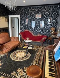 Victorian-style parlor interior with black damask wallpaper, red velvet chaise lounge and patterned settee, upright piano, crystal chandelier, and ornate floral rug