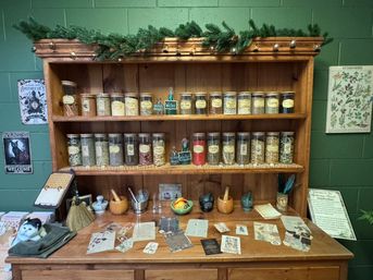 Cozy apothecary display: wooden shelves lined with labeled glass jars of dried herbs, mortar-and-pestles, recipe cards and botanical prints on a green wall.