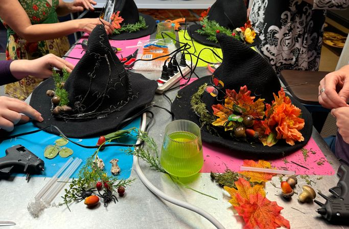 Hands decorating black witch hats at an indoor Halloween craft table with autumn leaves, orange flowers, moss, acorns, glue guns and craft supplies.