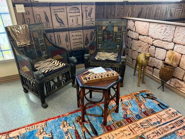 Egyptian-themed room with two ornate carved wooden chairs featuring cat-shaped armrests and zebra cushions, octagonal table with a wooden board game, hieroglyph wall panels, colorful Egyptian rug and camel statue.