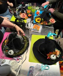 Top-down view of a DIY Halloween craft workshop table with black witch hats decorated with faux pumpkins, sunflowers, silk flowers, moss and spider webs, surrounded by hot glue guns, scissors and other craft supplies.