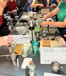 Hands-on DIY perfume workshop scene: people mixing essential oils and clear liquids on a stainless-steel worktable with glass bottles, droppers, measuring cups, small jars and printed recipe sheets.
