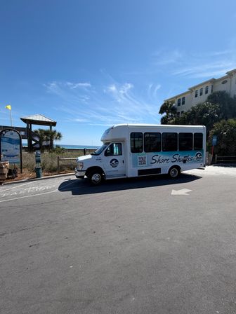 White beach shuttle parked by a public beach access and wooden boardwalk, with palm trees, sandy dunes and turquoise ocean under a bright blue sky