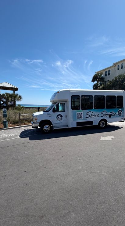 White beach shuttle parked by a public beach access and wooden boardwalk, with palm trees, sandy dunes and turquoise ocean under a bright blue sky