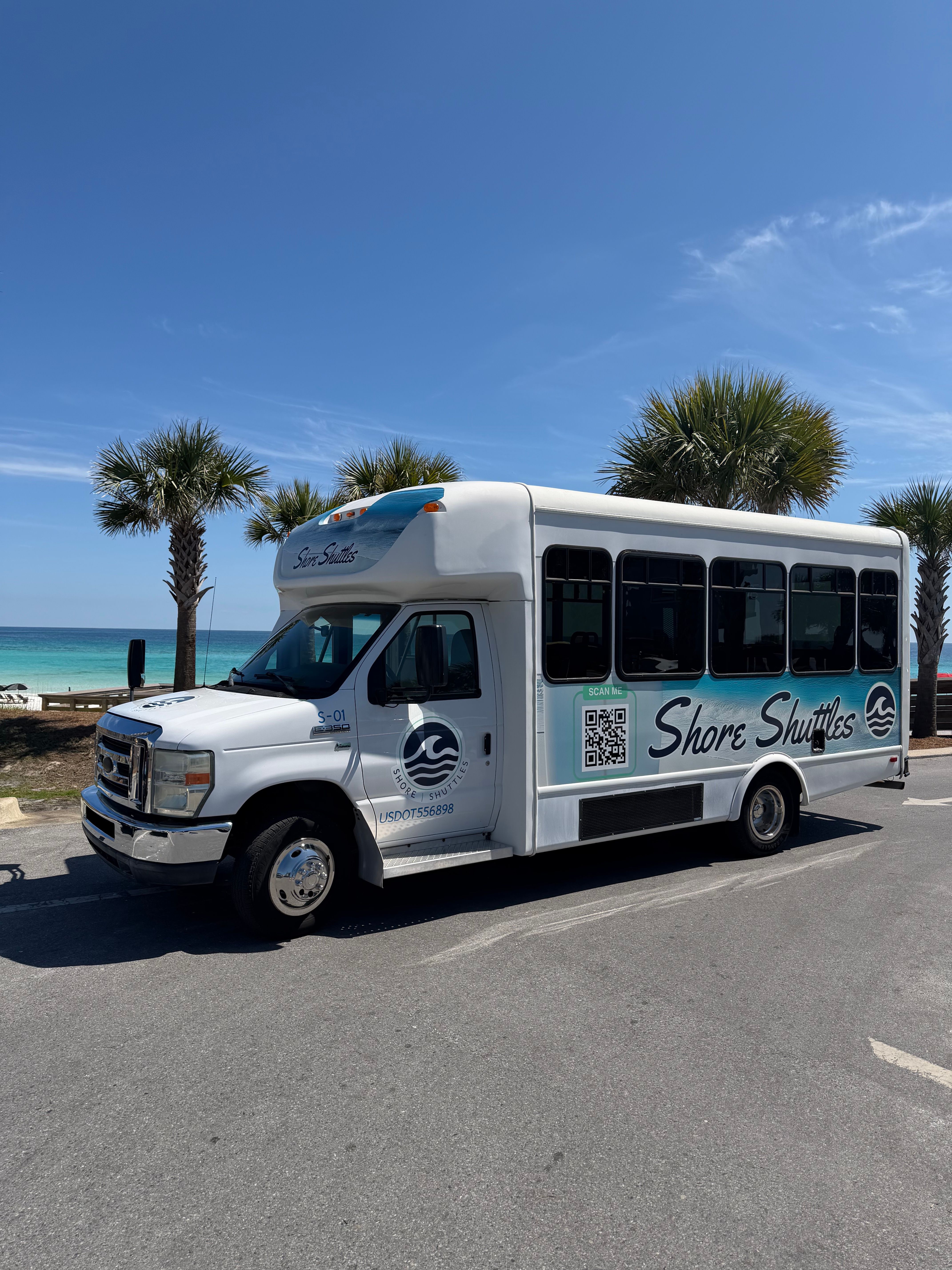 White shuttle bus parked at a sunny beachfront with palm trees, turquoise ocean and a clear blue sky.