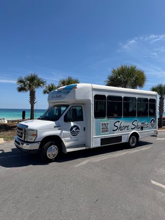 White shuttle bus parked at a sunny beachfront with palm trees, turquoise ocean and a clear blue sky.